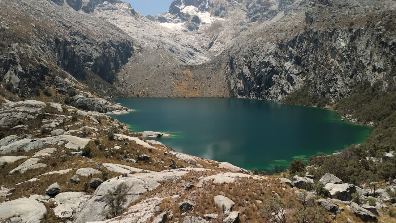 Drone slowly ascends with upward tilt, capturing the vivid turquoise waters of Laguna Churup and the surrounding rocky Andes in Huascarán National Park, Peru