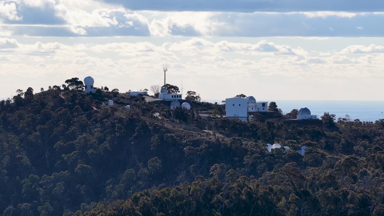 Drone camera slowly pans across a mountaintop observatory complex surrounded by dense forest under partly cloudy daylight, revealing multiple domed structures and rugged terrain