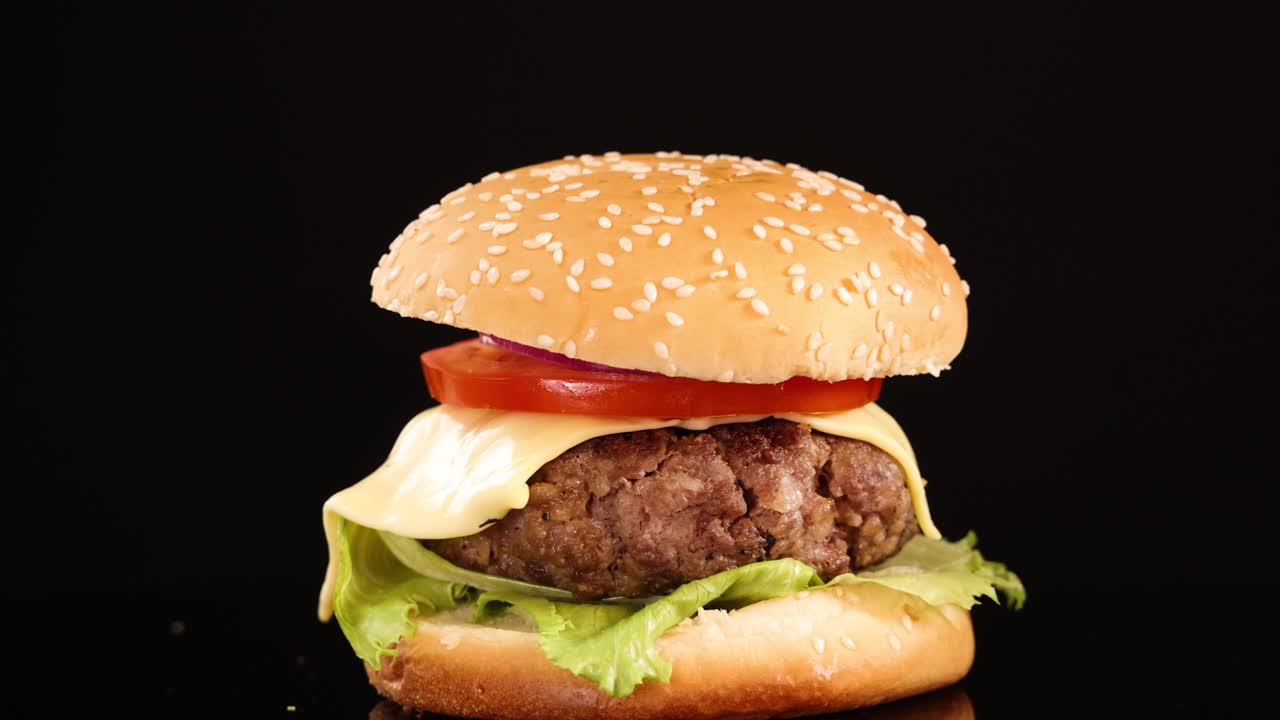 A juicy beef burger with cheese, lettuce, tomato, and onion rotates smoothly against a black background under bright, even studio lighting