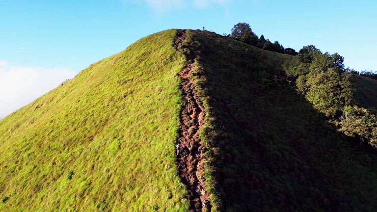 Un escursionista solitario che scala un ripido sentiero sterrato su una lussureggiante cresta montuosa tropicale verde verso la cima in una giornata di sole splendente, riprese aeree con drone