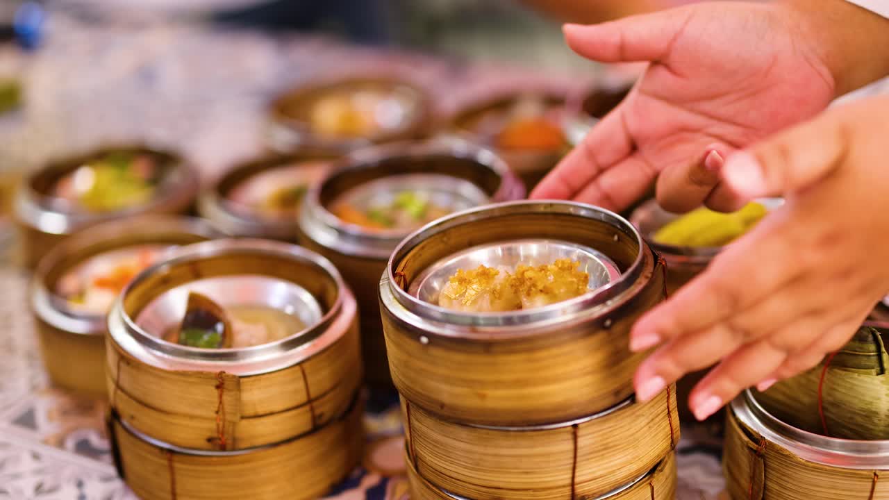 Hands choose dim sum from bamboo steamers in a vibrant market. Warm lighting enhances the lively, bustling atmosphere