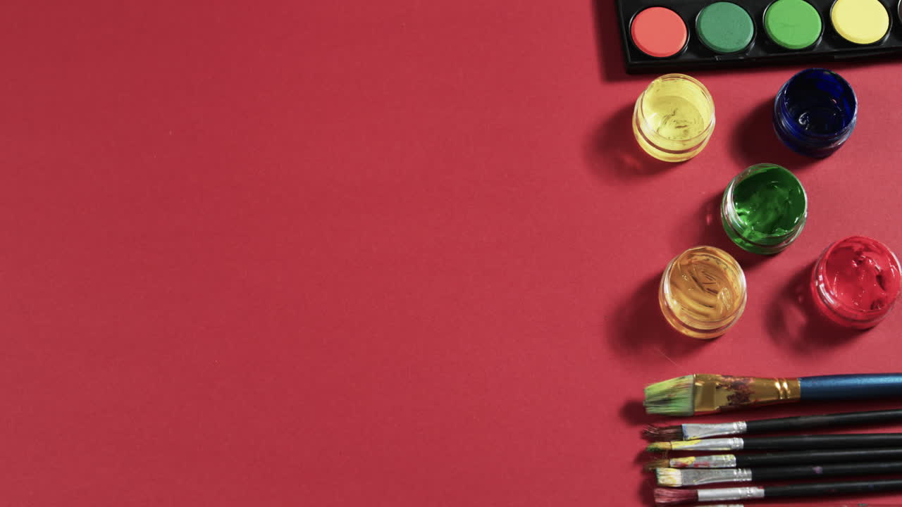 vista aérea de pinturas y pinceles dispuestos sobre un fondo rojo con espacio de copia, en cámara lenta