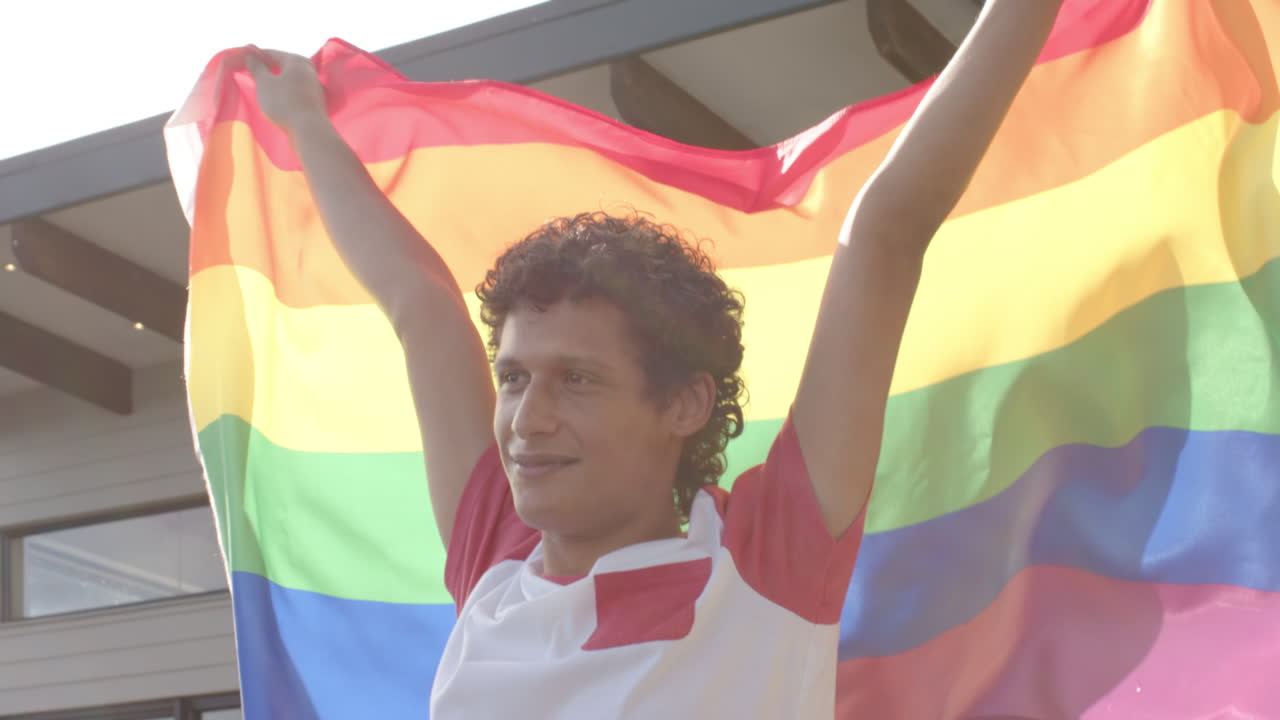 Uomo birazziale felice che tiene la bandiera arcobaleno LGBT sul balcone di casa, al rallentatore