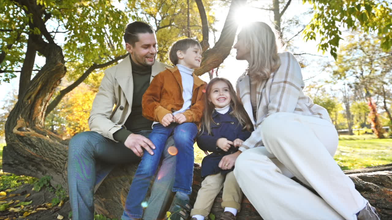 Happy family in an autumn park. Mother, father, son and daughter sitting on a tree trunk, mom kissing her son, yellowed trees around. Slow motion