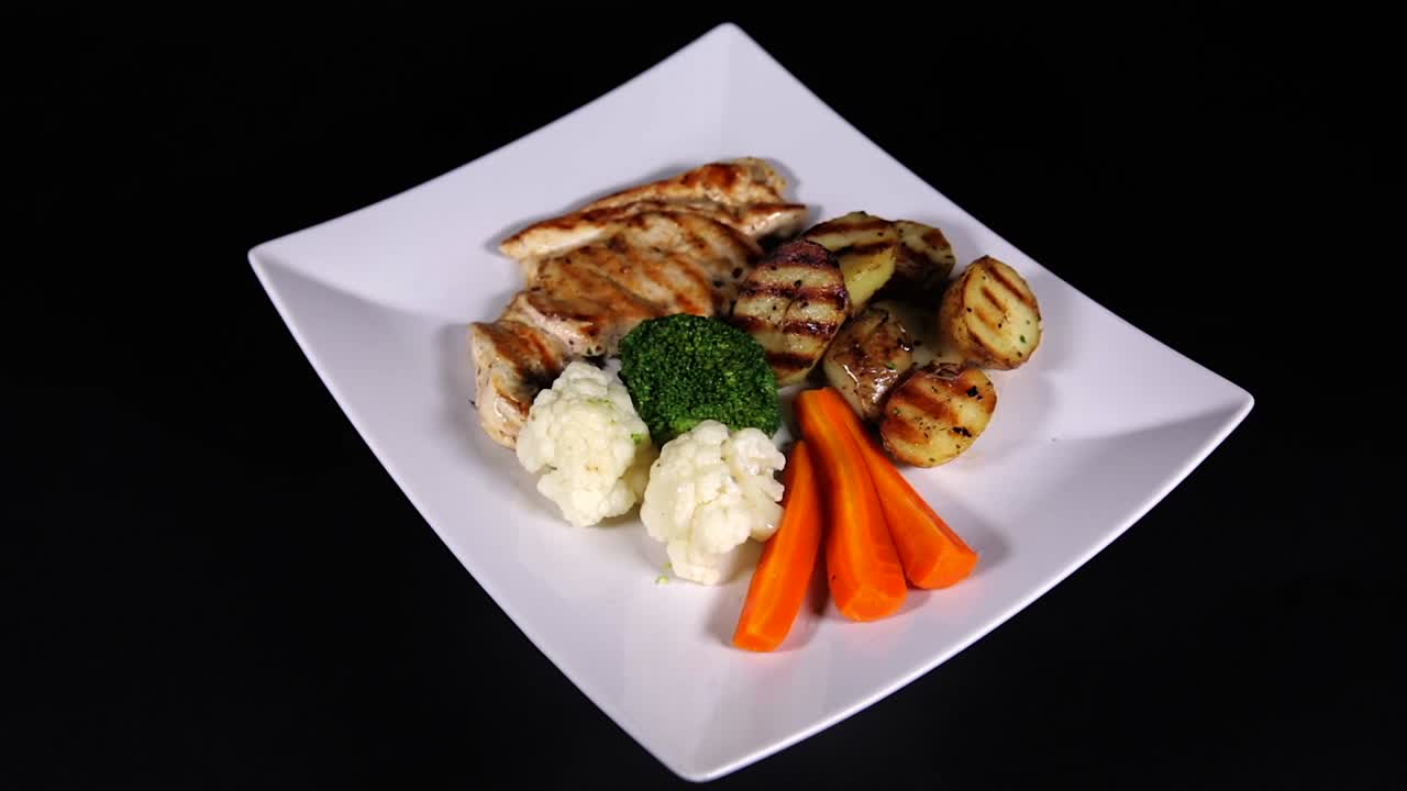 Spinning view of grilled chicken, veggies, and roasted potatoes on square plate.