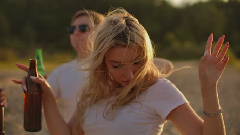 a blonde girl with bright makeup and long hair which is flying on the wind enjoys dancing and party time with beer and friends. this is carefree time at sunset on the river coast.