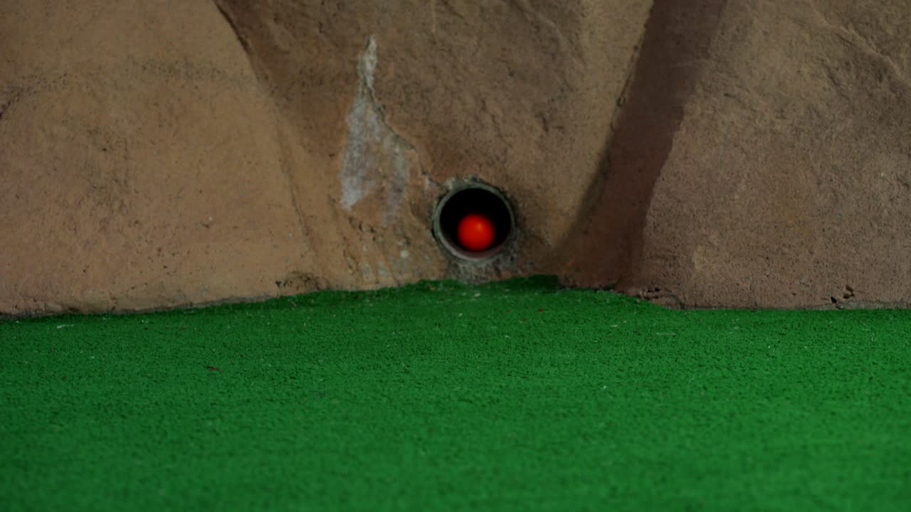 An orange mini golf ball exits a pipe in a wall and rolls over and misses the hole