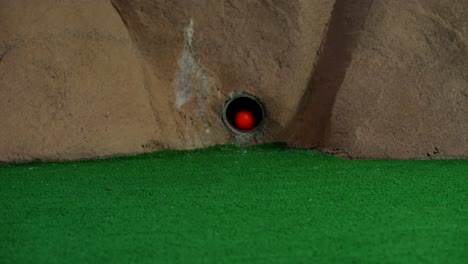 An-orange-mini-golf-ball-exits-a-pipe-in-a-wall-and-rolls-over-and-misses-the-hole