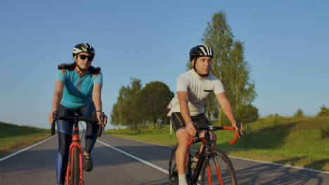 Two-professional-road-cyclists-ride-their-bikes-on-a-hill.-Hand-held-shot-of-two-strong-cyclists-female-and-male-on-their-training-on-a-warm-but-windy-summer-day