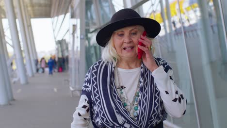 senior tourist grandmother woman walking on international airport hall, using mobile phone, talking