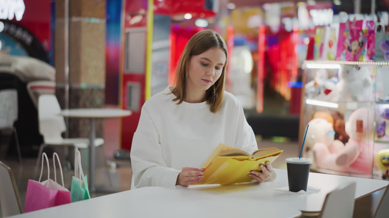mujer sentada en un centro comercial leyendo un libro amarillo, con taza de café y bolsas de compras de colores en la mesa, rodeada de iluminación suave, decoración vibrante y fondo borroso de juguetes y compradores