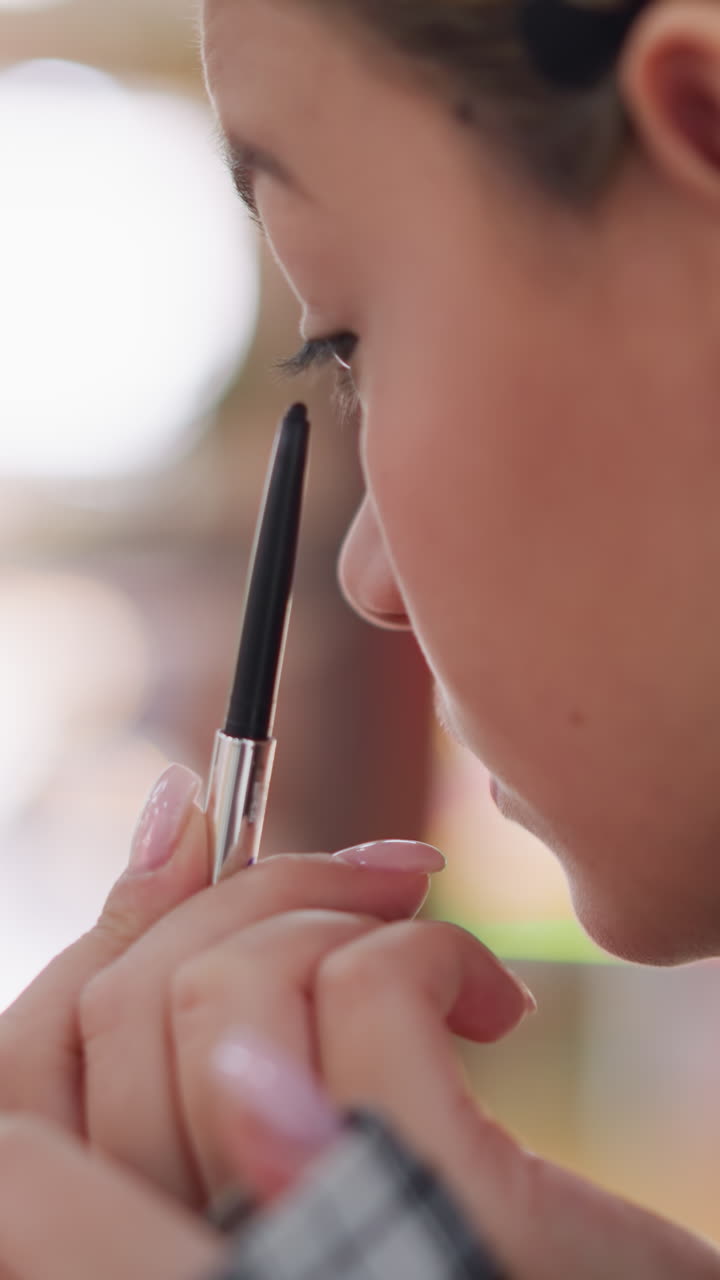 Primer plano lateral del perfil de una mujer sin especificar aplicándose un lápiz de ojos con luz tenue, modelo preparándose en el backstage con movimientos cuidadosos de las manos y uñas arregladas, ambiente íntimo de salón que muestra concentración