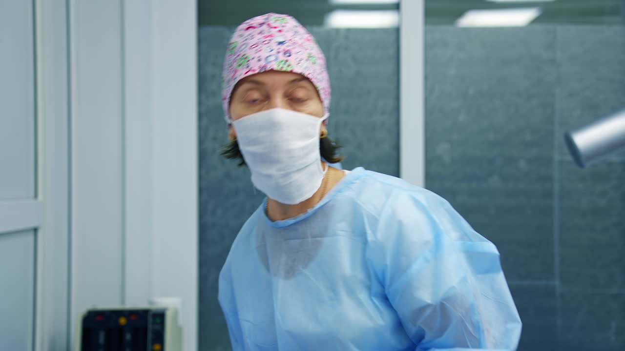 Professional ophthalmologist discusses surgery operation to the camera. Woman medic in scrubs and mask stands on modern equipment background.