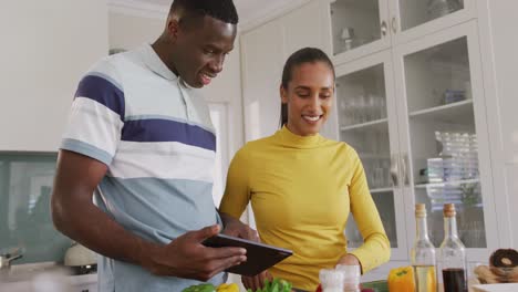 Video-of-happy-diverse-couple-using-recipe-on-tablet-preparing-food-together-in-kitchen