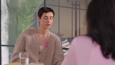 two women wearing pink breast cancer awareness ribbons meeting and talking at therapy support group for cancer treatment patients