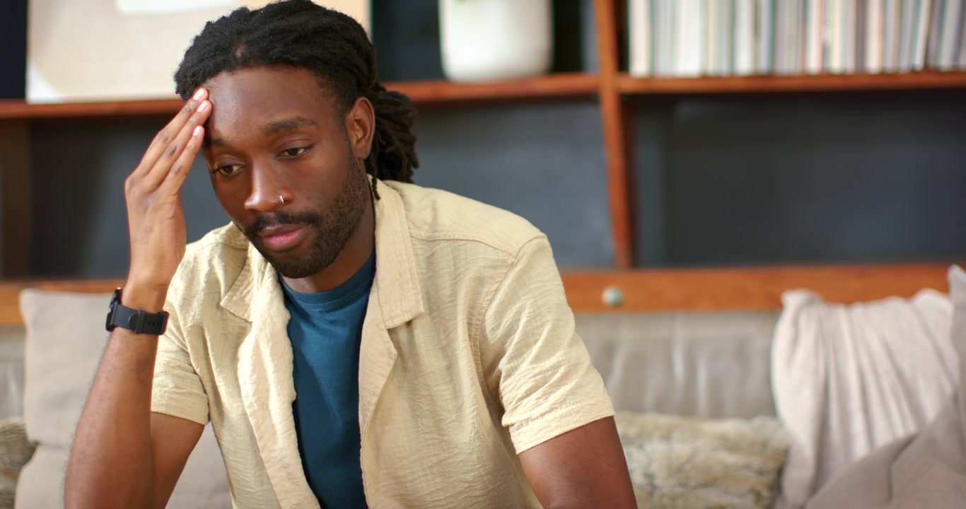 African male sitting on sofa at home after prompt, preparing response gesturing and touching temple