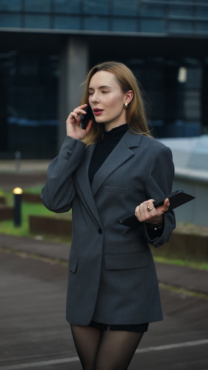Happy young woman in a blazer smiles during a phone conversation while holding a tablet on an office terrace - tarallax shot, slow motion