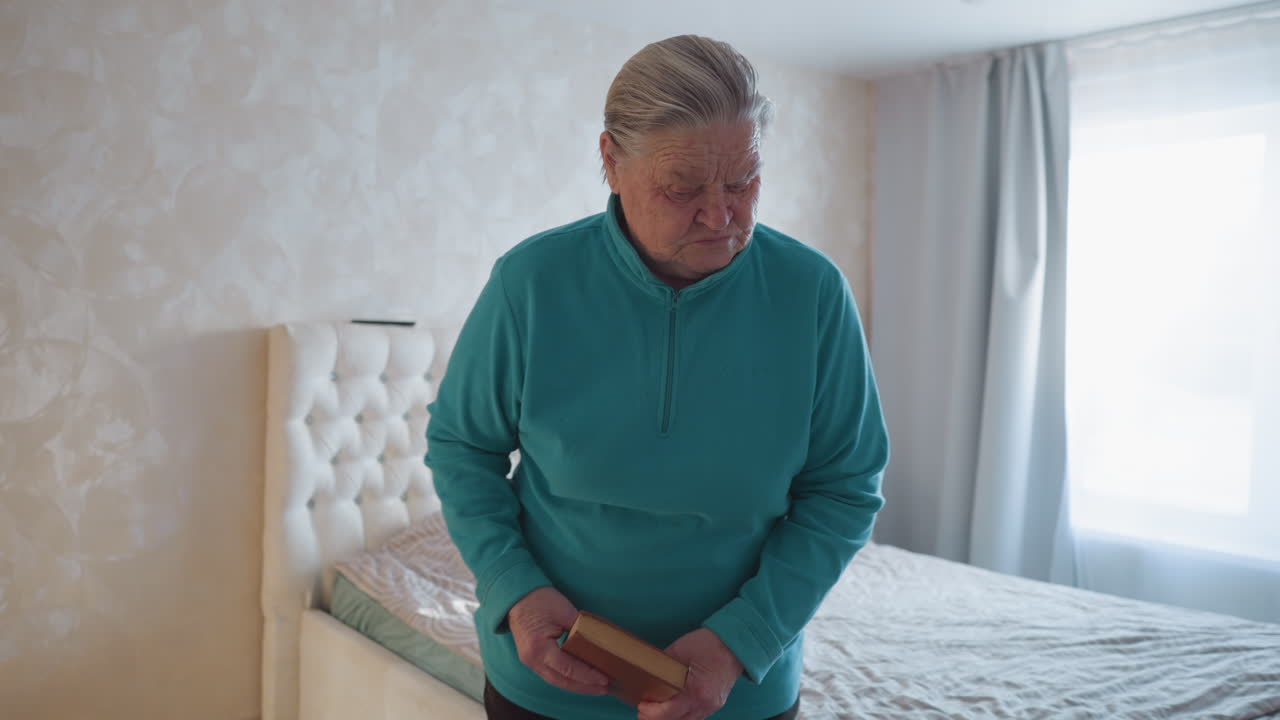 Elderly woman walking across room holding book, wearing teal sweater, walking past cozy bedroom with natural sunlight streaming in, calm indoor atmosphere, minimalistic decor, and peaceful environment