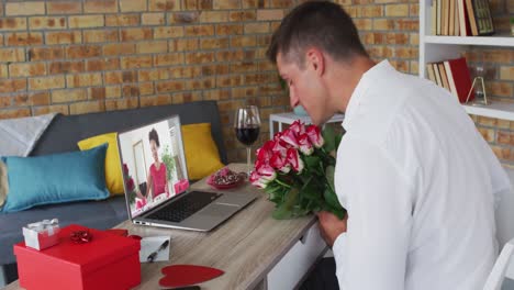 Diverse-couple-on-a-valentines-date-video-call-both-holding-flowers-man-blowing-kiss-to-woman-on-scr