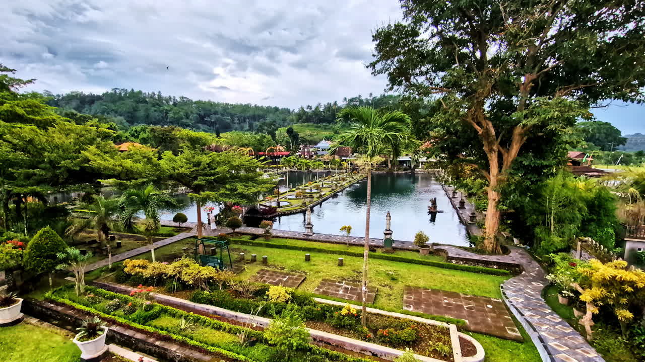 Slow rightward panning shot of the beautiful Tirta Gangga Water Palace in Bali, featuring lush gardens, stone statues, and reflective pools surrounded by tropical greenery