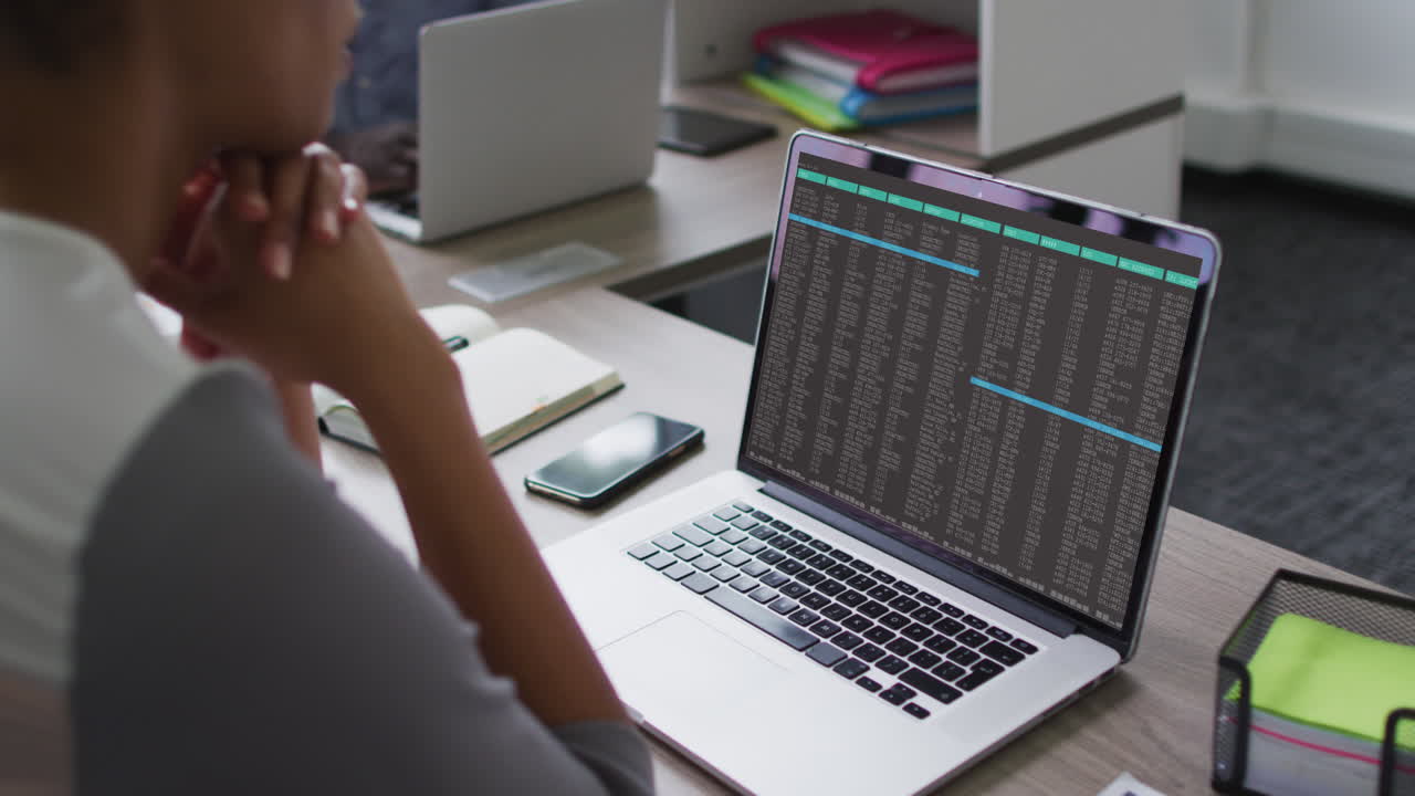 Biracial woman sitting at desk watching coding data processing on laptop screen