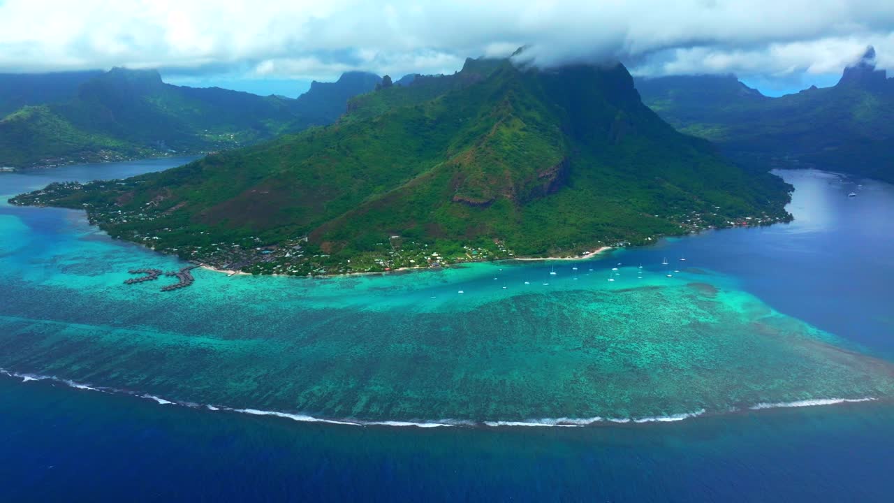 Luftfoto af Moorea, Fransk Polynesien, med Opunohu-bugten, en bred koralrevshylde og levende turkis vand, der buer langs øen under skybeklædte grønne bjerge, panorerer opad