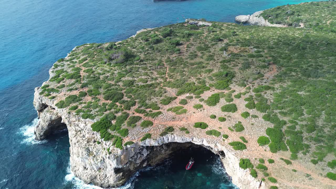 4K drone footage of Mallorca’s rocky shoreline with large sea caves in the cliffs. A red diving boat is anchored inside the biggest cave, surrounded by crystal blue water and green vegetation