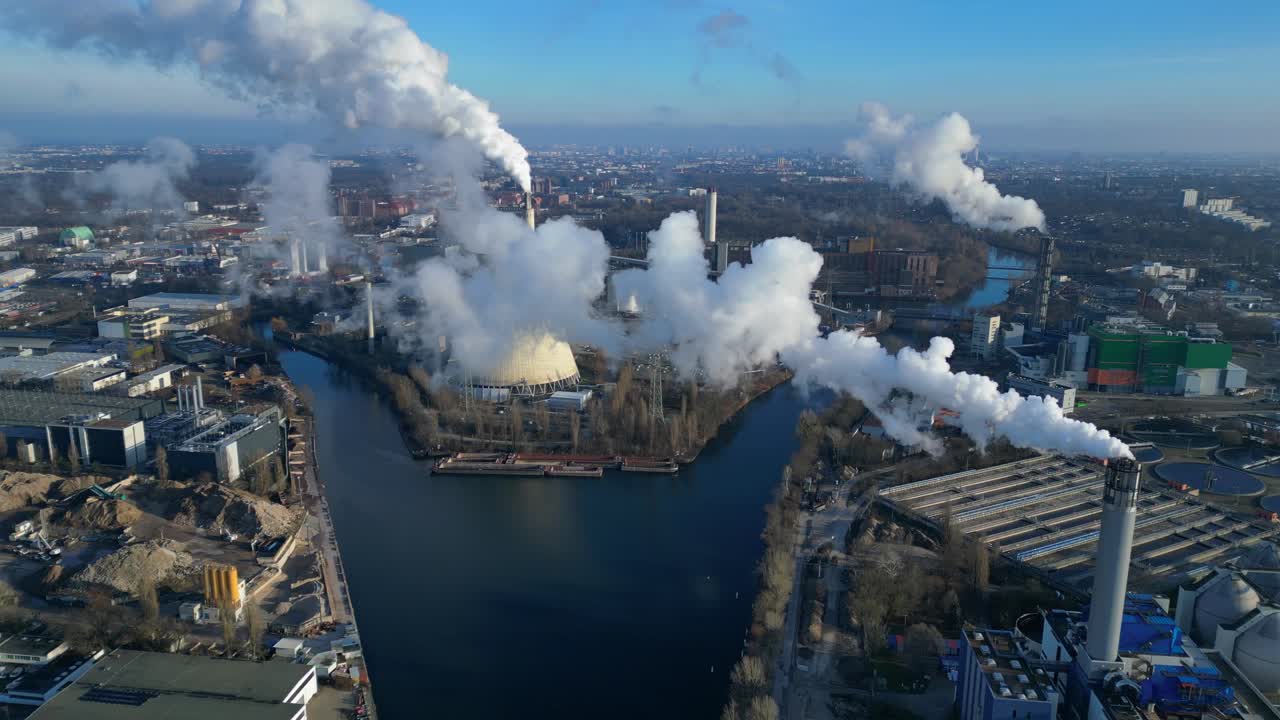 Aerial view of a thermal power plant emitting smoke on a sunny day, with a river and a city in the background. Stunning aerial view flight drone top down Above view