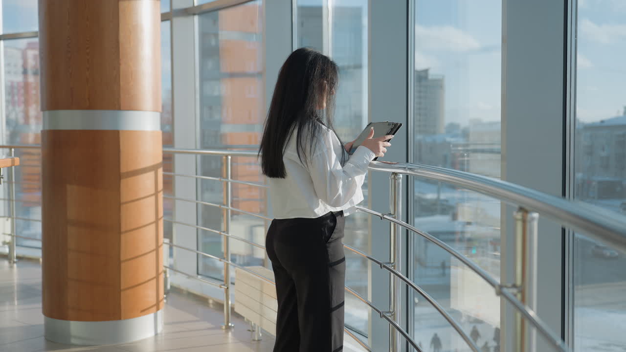 Rear view of businesswoman holding tablet walking toward chrome railing in bright office hallway with floor to ceiling windows offering view of winter cityscape under blue sky