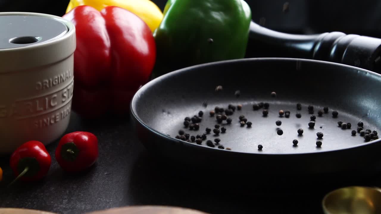 Slow motion black peppercorns being poured into a black bowl. In the background is garlic, red and green peppers and small red peppers. Making a vibrant Cajun Spice Blend with many herbs and spices.