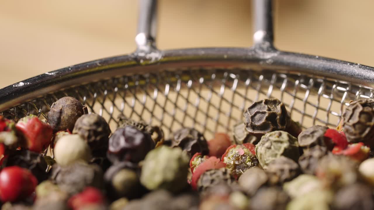 Camera focuses on mix of colored dry peppercorns. Close up of pink, black and white peppercorns lying in sieve on the kitchen table. Spices and condiment. Falling seeds of bitter peas in slow motion