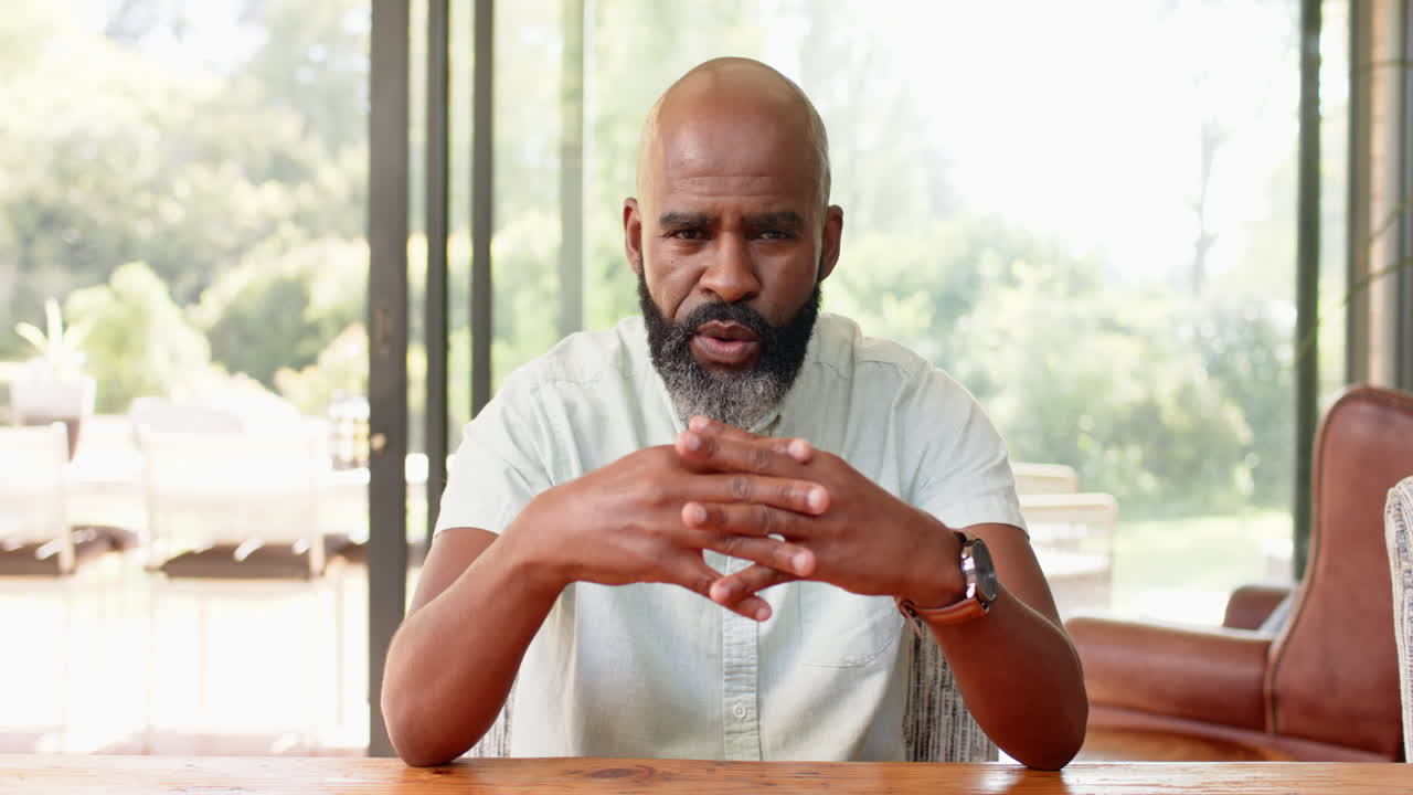 Senior man explaining and gesturing while on video call at wooden table indoors