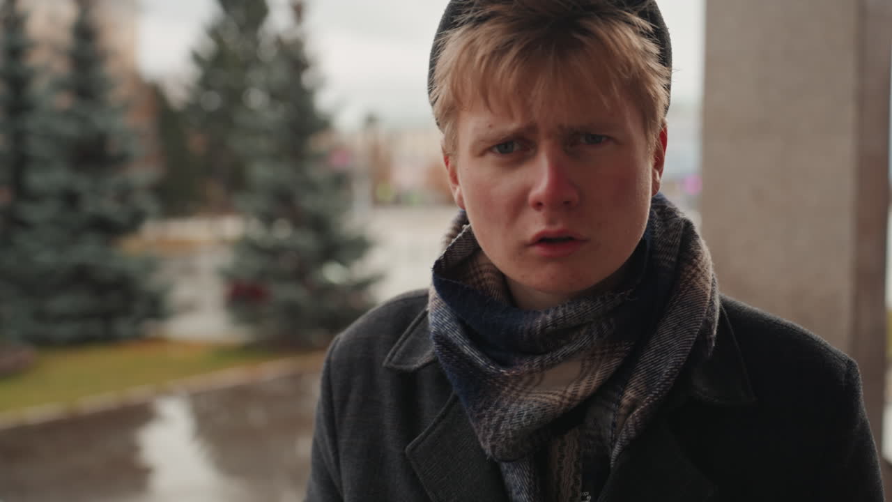 White creator removes sunglasses on balcony, moody closeup portrait of white man with scarf and coat, rainy urban backdrop with pine trees and reflective puddles, introspective expression