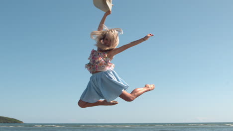 attractive blonde holding straw hat jumping on the beach