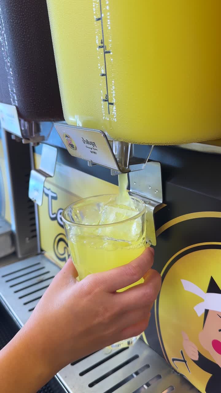 Una mano llena un vaso con hielo usando una máquina amarilla de bebidas heladas en Bangkok