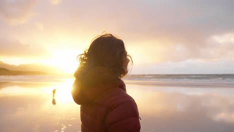 SLOW-MOTION,-a-beautiful-young-woman-admiring-the-view-and-smiling,-with-wind-in-her-hair,-on-the-Flakstad-beach-in-front-of-a-gorgeous-golden-sunset,-Lofoten-Islands,-Norway