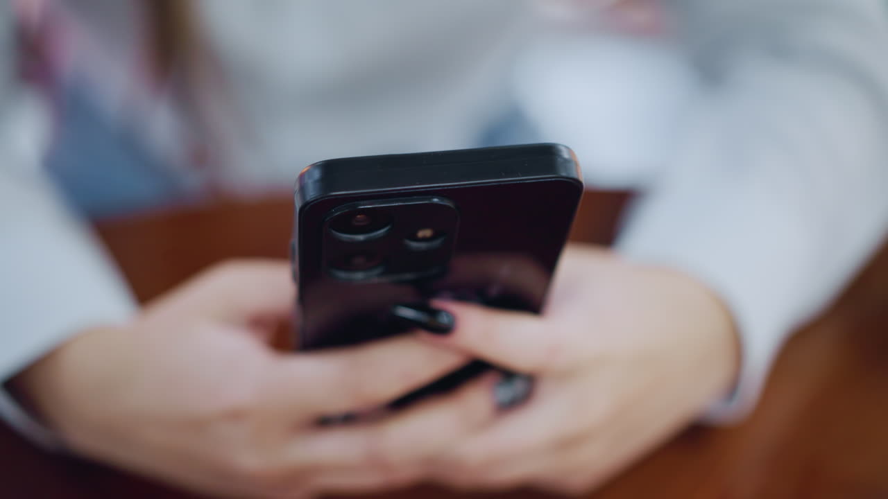 vista borrosa de la mano de la dama con uñas pintadas interactuando con el teléfono inteligente colocado en una mesa de madera, centrándose en la tecnología moderna y la moda en un entorno suave y vibrante con un fondo borroso
