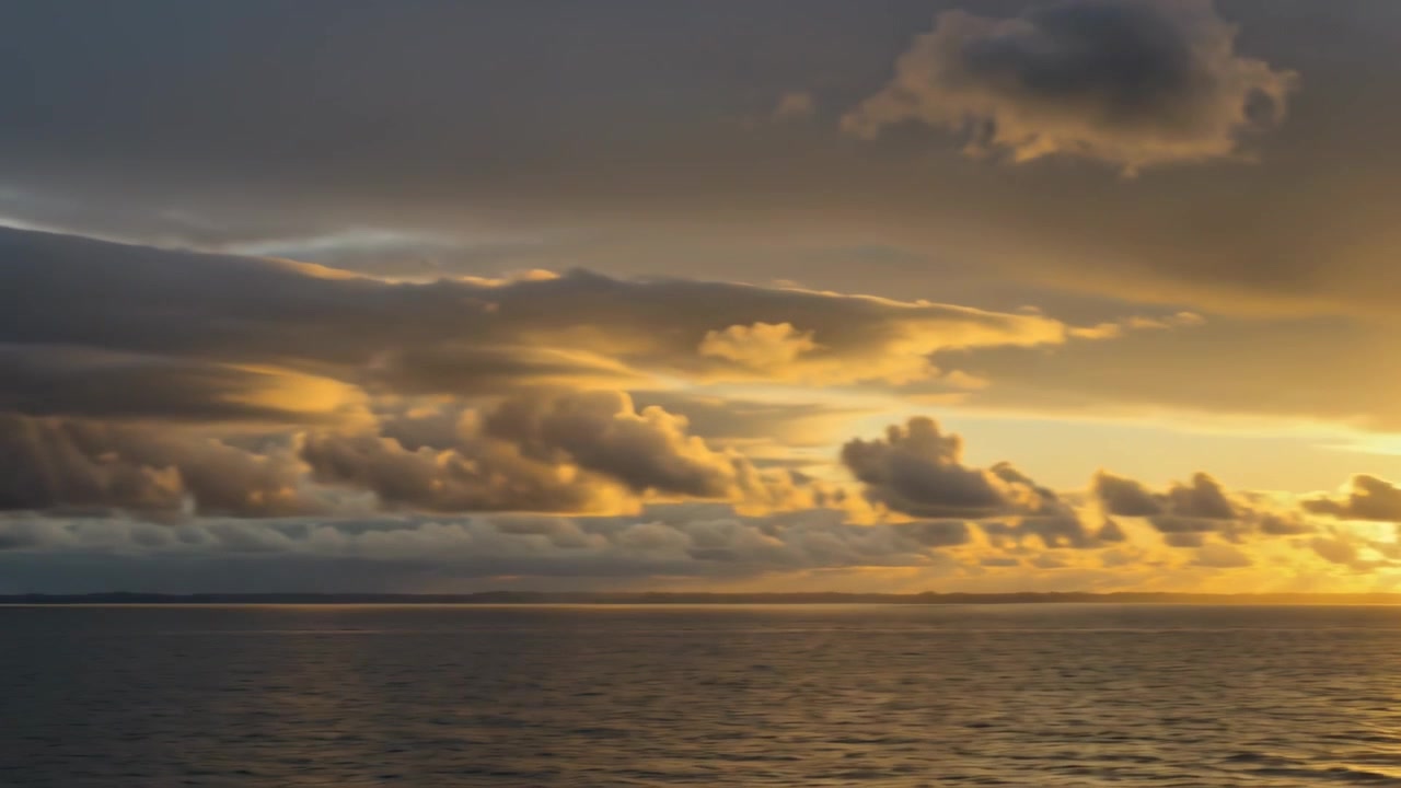 Die aufgehende Sonne färbt eine geschichtete Wolkenbank über dem ruhigen Ozean an der Küste und wirft eine goldene Reflexion.