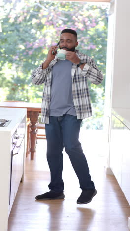 african american man in kitchen talks on phone, sips coffee, vertical slow-mo video