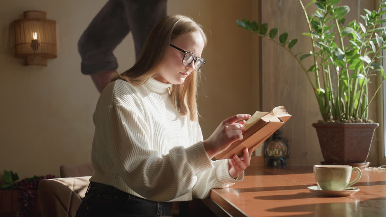 Reflective girl in glasses reading book near window with warm natural light, blur background showing wall art, indoor plant, and mug on table
