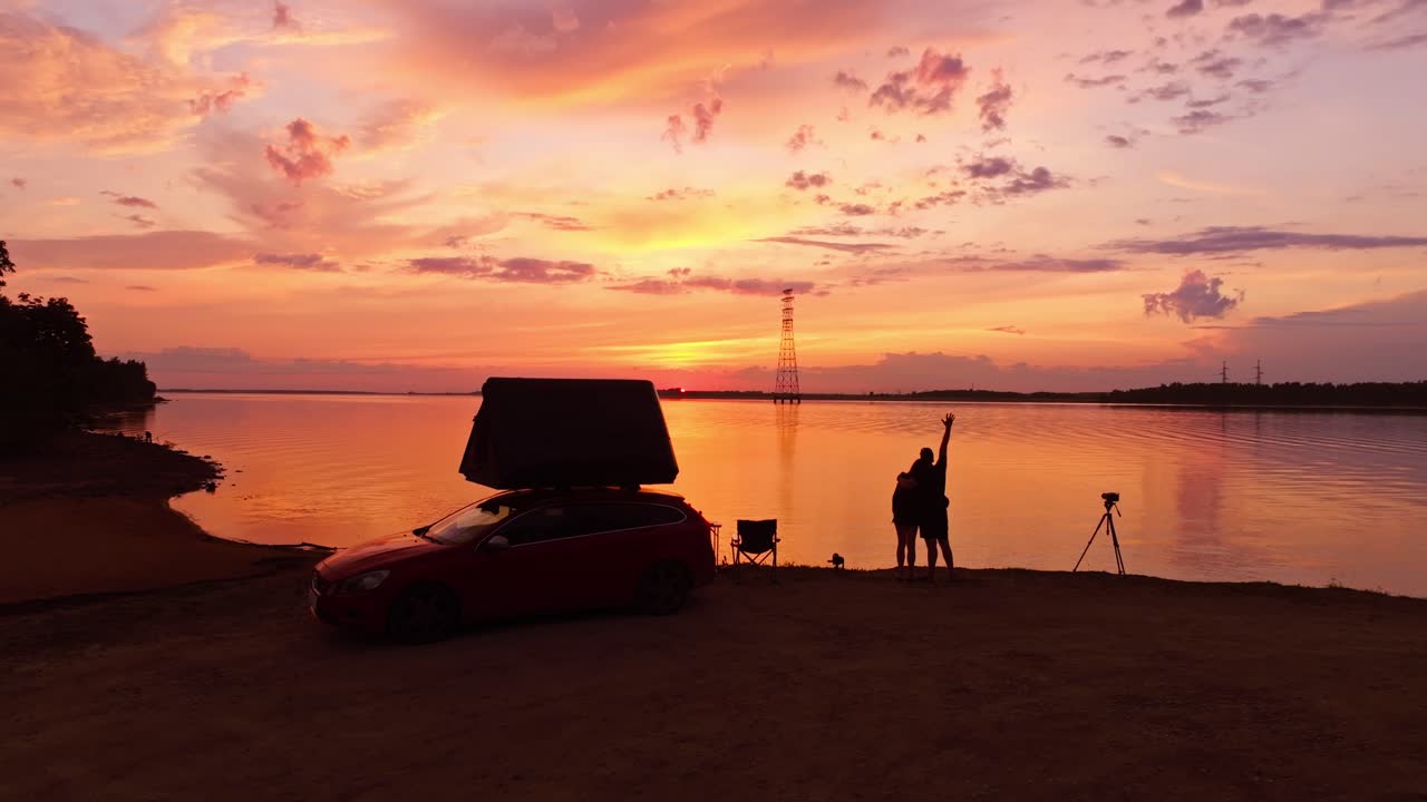 Golden lake sunset with couple hugging by car tent, camera on tripod nearby