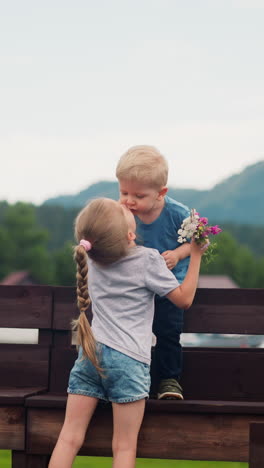 little girl with plait kisses younger brother standing on wooden bench to thank for flower bouquet gift at resort in spring slow motion