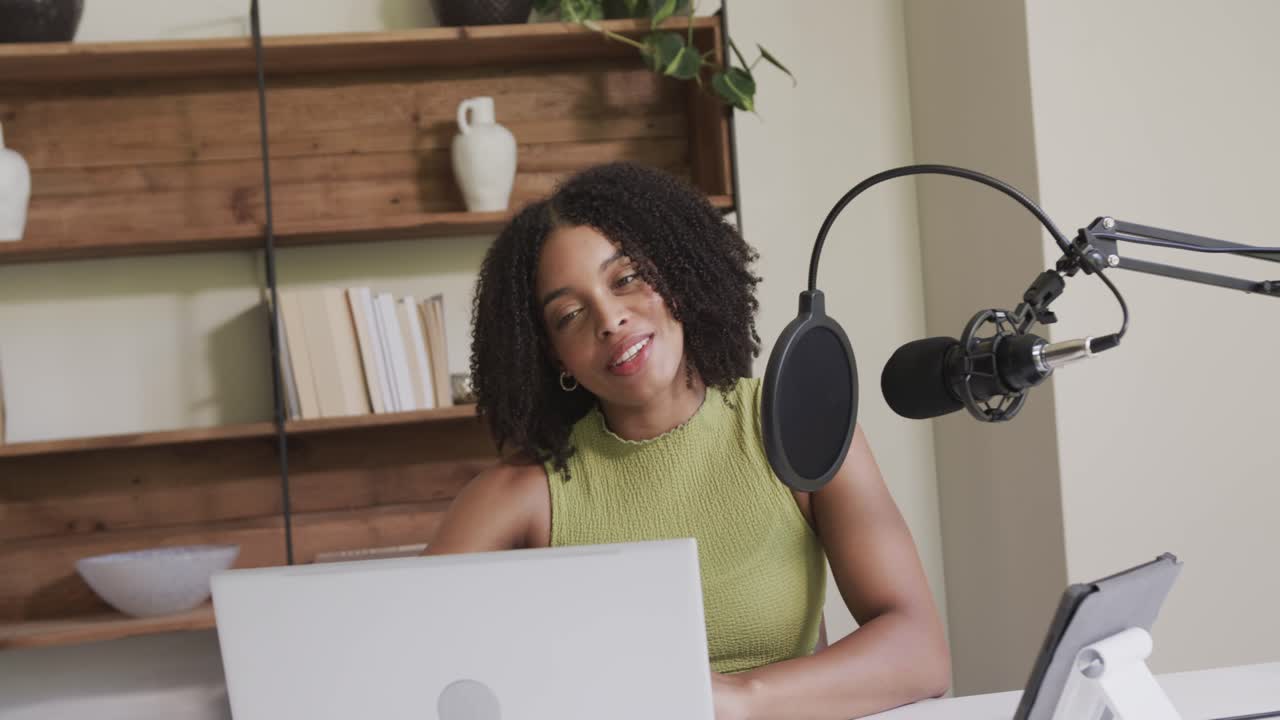 African American woman in studio in green top, going live, talking into mic, reading laptop or table