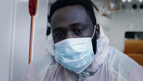 black emergency service worker taking off face shield african american paramedic in mask taking off face shield and hazmat suit hood while looking at camera in ambulance during pandemic