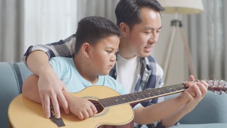 close up of asian father teaching his son about guitar chords on sofa at home