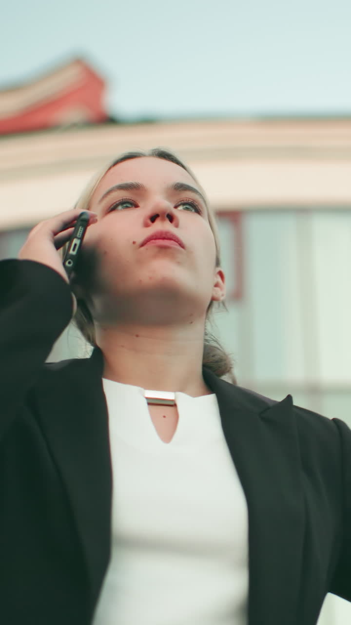 Fed up lady in professional outfit speaks on phone with tired expression in urban setting, modern building slightly blur in background
