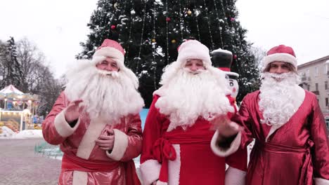 three santa clauses on the background of a christmas tree in the city center three santa clauses on the background of a christmas tree in the city center group of santa clauses waving at the camera christmas and new year concept