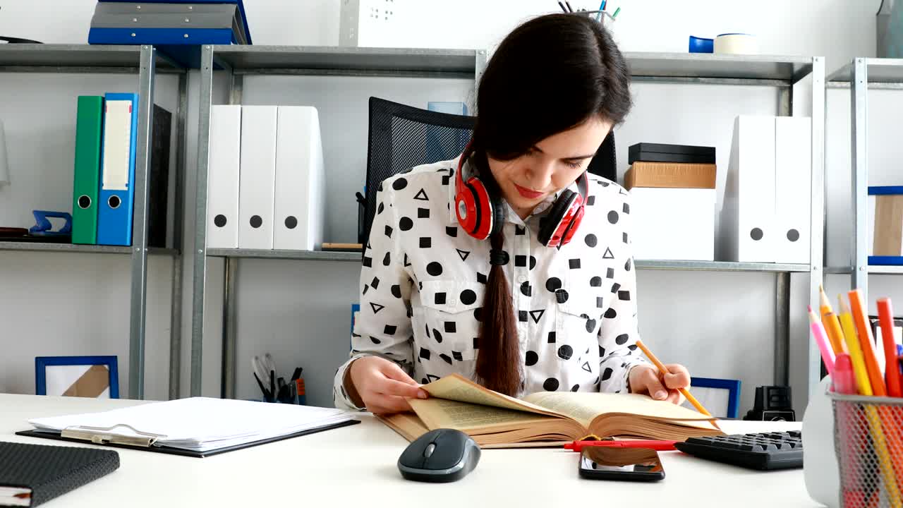 woman with red headphones on shoulders thumbing book and writing in pencil on paper
