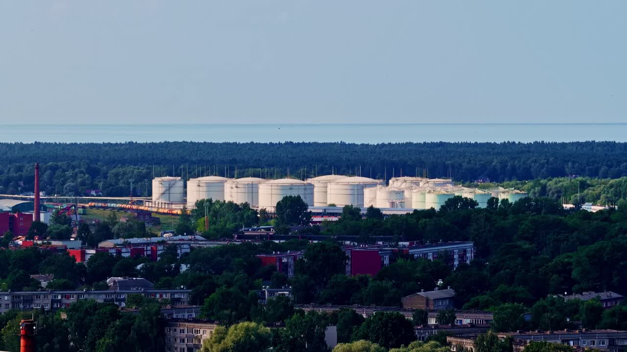 Aerial establishing of large white oil storage tanks near the coastline, framed by dense green forest and city outskirts under a clear blue summer sky