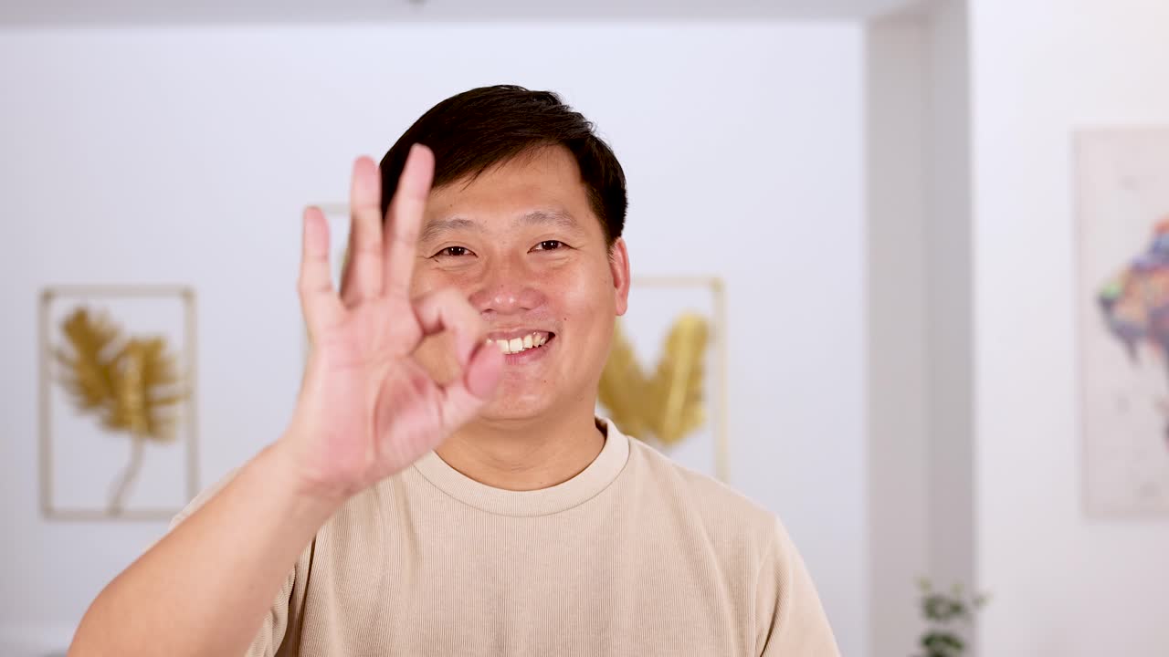 Asian man smiles and raises his hand to make an okay gesture in a bright, modern indoor setting with soft, even lighting
